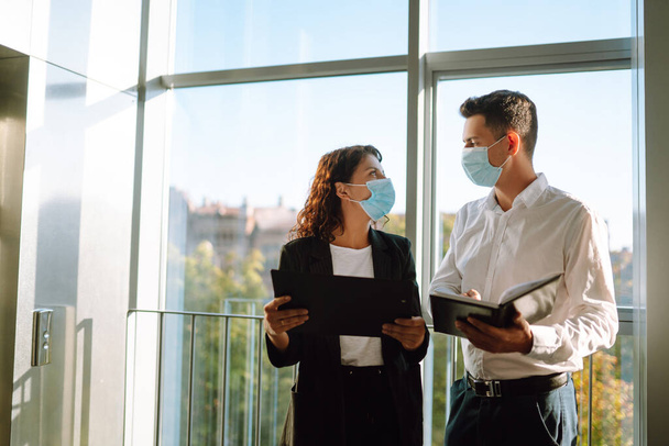 Two colleagues in protective face masks in modern office discussing together work issues standing near elevator. Business People back at work in office after quarantine. Covid-19. - Foto, Imagem