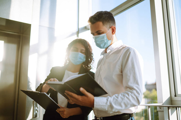 Two colleagues in protective face masks in modern office discussing together work issues standing near elevator. Business People back at work in office after quarantine. Covid-19. - 写真・画像
