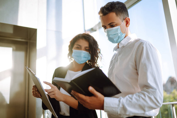 Two colleagues in protective face masks in modern office discussing together work issues standing near elevator. Business People back at work in office after quarantine. Covid-19. - Photo, Image