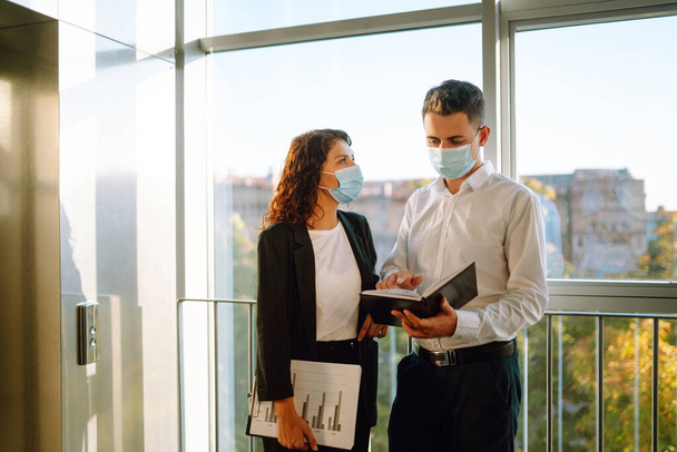 Two colleagues in protective face masks in modern office discussing together work issues standing near elevator. Business People back at work in office after quarantine. Covid-19. - Φωτογραφία, εικόνα