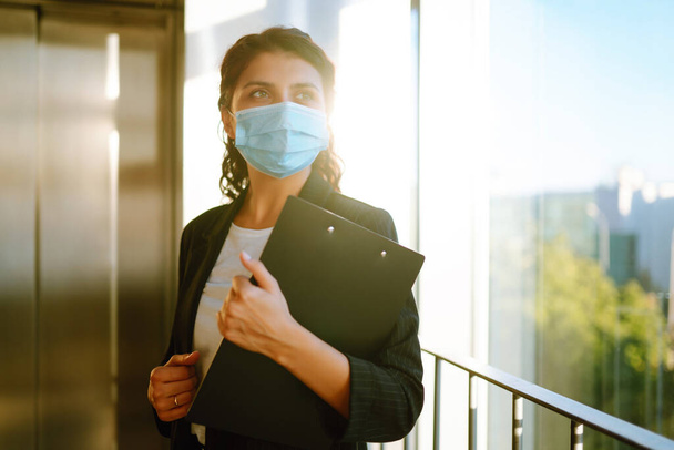 Portrait of businesswoman in protective face mask during virus epidemic while standing near elevator at modern office. Office manager back at work in office after quarantine. Covid-19. - Valokuva, kuva