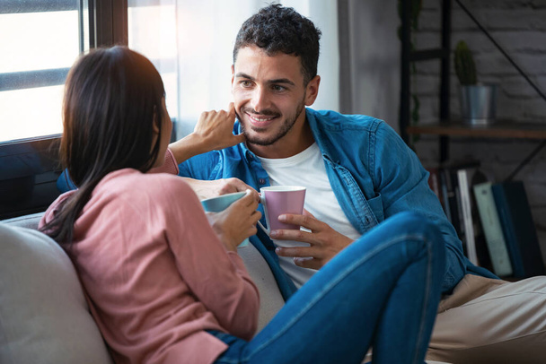 Shot of lovely couple holding coffee cups while looking at each other and smiling sitting on couch at home. - Φωτογραφία, εικόνα