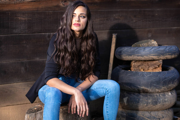 Portrait of a beautiful brunette girl in jeans, sitting next to a pile of old tires - Foto, afbeelding