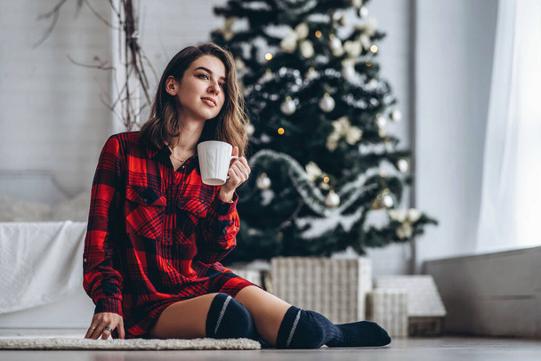 Pretty brunette girl sitting on the floor in red shirt and warm socks with cup of coffee and christmas tree behind - Фото, изображение