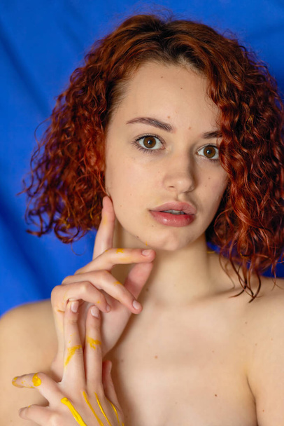 redhead curly woman hands in yellow paint looks at the camera on a blue background. Concept photography for art or women's blog - Fotografie, Obrázek