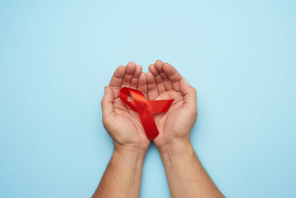 mains masculines tient un ruban rouge - un symbole de la lutte contre la maladie SIDA et vascularite, fond bleu, vue du dessus - Photo, image