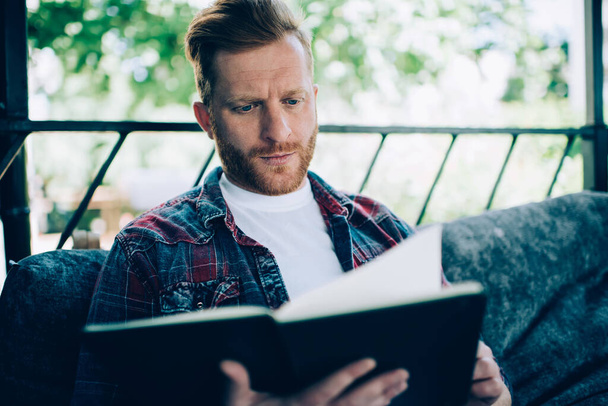 Focused male startupper in casual outfit learning with book while having break at outdoor cafe veranda with trees on blurred background - Photo, image