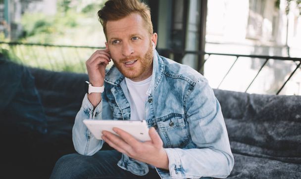 Bearded redhead casual male in denim jacket sitting on couch in city with tablet and wireless headphones and looking away - Foto, Bild