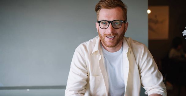 Smiling positive stylish ginger male freelancer in eyeglasses texting on smartphone while sitting alone at cafe table looking at camera - Фото, изображение