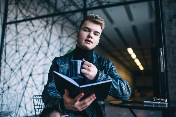 Low angle of young man in trendy leather jacket enjoying cup of coffee and looking at camera while reading book in cafe - Foto, Imagen