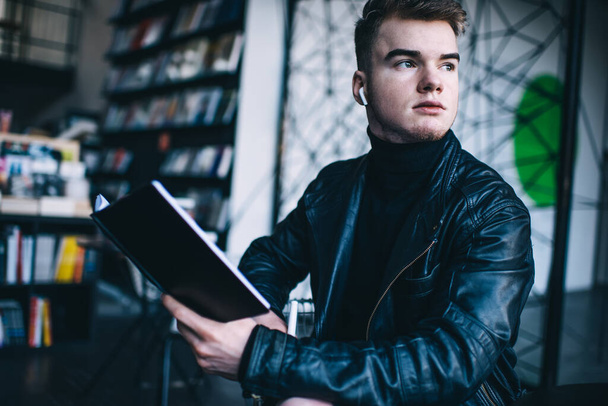 Serious young male in trendy black leather jacket with true wireless earbuds looking away while reading book in modern library - 写真・画像