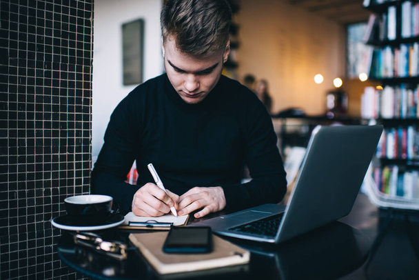 Concentrated male employee sitting at desk in cozy workspace and writing in clipboard while browsing laptop and drinking cup of coffee - Foto, immagini
