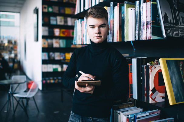 Thoughtful young male in casual wear with modern haircut standing with pen and notepad near bookcase behind table and chairs in library while looking at camera - Foto, immagini