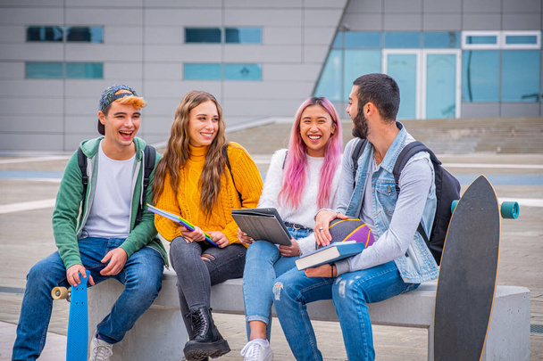 Monietniset opiskelijat vitsailevat ja puhuvat istuen penkillä yhdessä ulkona yliopistossa - ryhmä onnellisia nuoria nuoria opiskelee kirjoja ja tabletteja - Valokuva, kuva