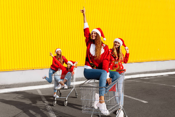 Happy young women friends in christmas sweaters and santa claus hats celebrate new year and ride trolleys against yellow wall background - Photo, Image