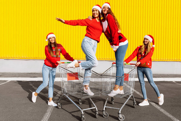 Group of four young women in Christmas sweaters and Santa Claus hats having fun on carts near a shopping center - Photo, Image