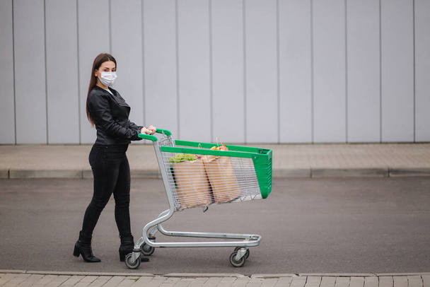 Mladá atraktivní žena chodící s nákupem vozíků ze supermarketu. Sociální distancování pro ochranu, obličejová maska, aby se zabránilo infekci. Nákup potravin během karantény Coronavirus Covid-19. Strana - Fotografie, Obrázek