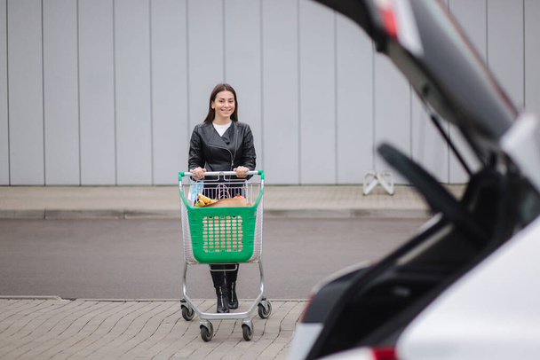 Młoda atrakcyjna kobieta spaceruje z wózkiem spożywczym z supermarketu do samochodu. - Zdjęcie, obraz