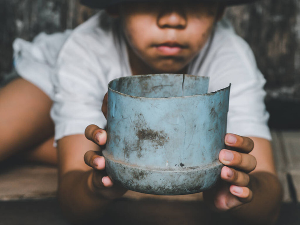 Child or poor homeless holding plastic bowl with hungry and asking help food donation from traveler on street at the city. Unidentified homeless child begging on street. Shelter concept. - 写真・画像