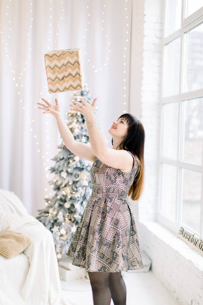 Christmas, x-mas, happiness, winter holidays. Beautiful charming woman in dress, having fun while celebrating Happy New Year and Christmas, posing in decorated room and throwing up present box - Fotoğraf, Görsel