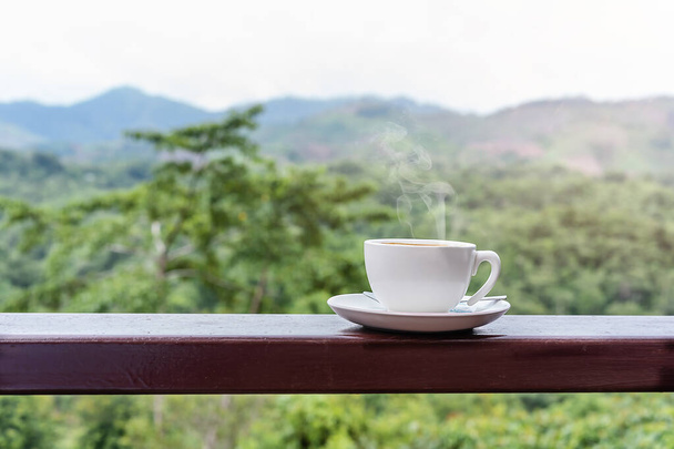 Lähikuva kuuma valkoinen kahvi kuppi puinen pöytä luonnonkaunis näkymä vuorelle aamulla auringonvalo ja kaunis vihreä luonto tausta kopioi tilaa tekstiä - Valokuva, kuva