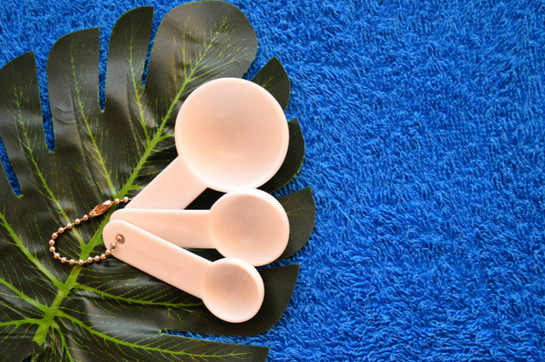 Белые пластиковые измерительные ложки лежат на пальмовом листке на синем фоне ткани - Фото, изображение