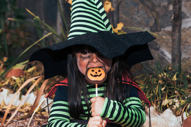 Boszorkánynak öltözött lány, cukorkát eszik, Halloweent ünnepli a kertben Jack O Lantern mellett. - Fotó, kép