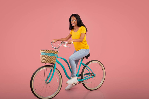 Full length portrait of beautiful African American woman riding vintage bicycle over pink studio background - Photo, Image