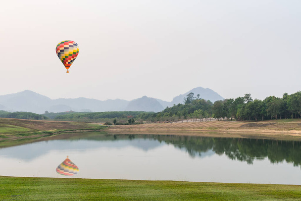 Photo of colorful aerostat balloon with basket floating in the air above lake with beautiful scenery behind it - Photo, Image