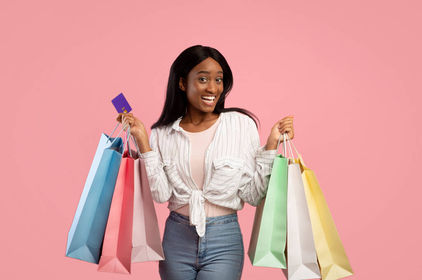 Concepto frenético de compras. Sonriente dama negra con bolsas de regalo y tarjetas de crédito comprando regalos navideños sobre fondo rosa - Foto, imagen