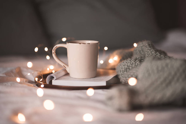 Ένα φλιτζάνι καφέ σε ανοιχτό βιβλίο με πλεκτό πουλόβερ στο κρεβάτι από κοντά. Καλημερα. Χειμερινές διακοπές.  - Φωτογραφία, εικόνα