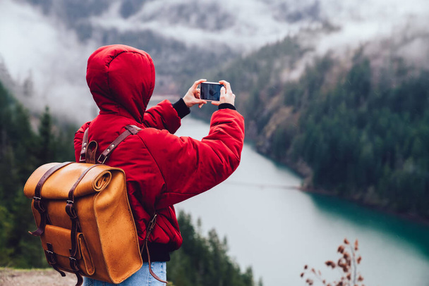 Back view of unrecognizable person in bright jacket photographing landscape on smartphone during trip in mountainous place on foggy day - Foto, imagen
