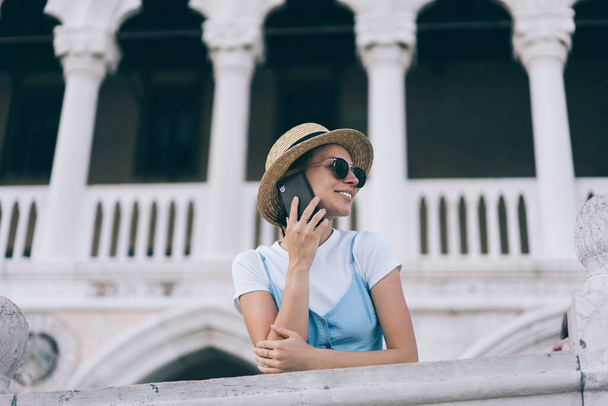 Low angle of smiling female spending time near aged monument while answering phone call during journey in Italy in summer - Φωτογραφία, εικόνα
