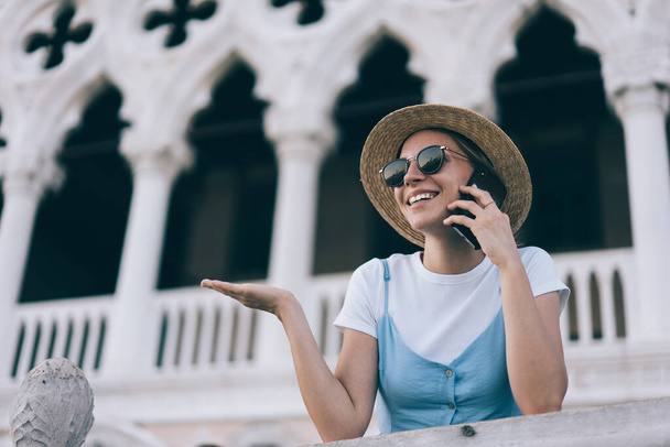 Low angle of cheerful female traveler in straw hat standing near historic building and chatting on cellphone during vacation in Venice - Photo, Image