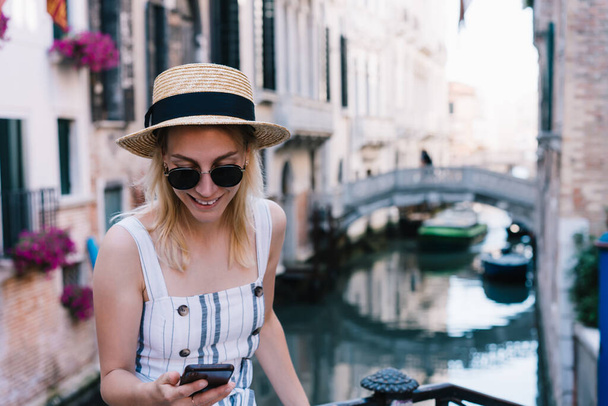 Content lady standing near vintage footbridge and holding smartphone while spending time in old city district near water channel in Venice - Photo, image