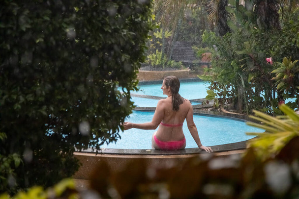 una chica en bikini se sienta al lado de una piscina al aire libre en un jardín tropical bajo la lluvia. - Foto, Imagen