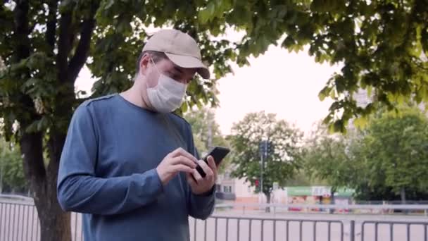Mężczyzna w masce ochronnej spacerujący ulicą, korzystający z internetu na smartfonie. - Materiał filmowy, wideo