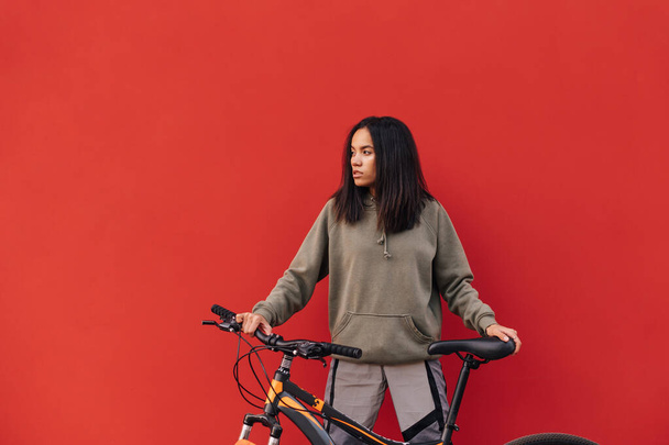 Belle femme hispanique en tenue de sport isolée avec un vélo sur fond rouge, regardant de côté l'espace de copie. Loisirs de vélo - Photo, image