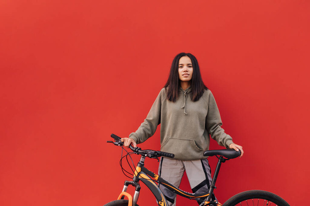 Позитивна іспаномовна жінка в стильному одязі стоїть з велосипедом в руках на червоному тлі і дивиться на камеру з посмішкою на її обличчі
. - Фото, зображення