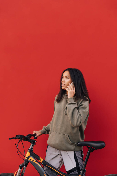 Portret van een aantrekkelijke Spaanse vrouw in casual kleding met fiets geïsoleerd op rode achtergrond, kijken opzij. Verticaal. - Foto, afbeelding