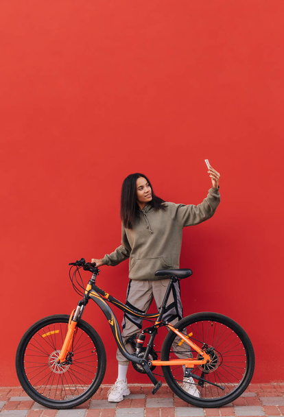 Positive woman sitting on a bicycle on a background of a red wall and taking a selfie on a smartphone. Isolated. Vertical - Photo, Image