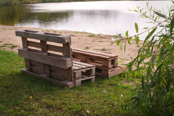 Зовнішні меблі з дерев'яних піддонів на березі тихого озера, оточеного деревами та зеленню на літньому сонці
 - Фото, зображення