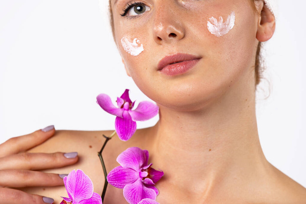 Beauty portrait. Girl with cream on her cheeks, making the skin softer and more hydrated. In hands with an orchid. Skin care concept. High quality photo - Photo, Image