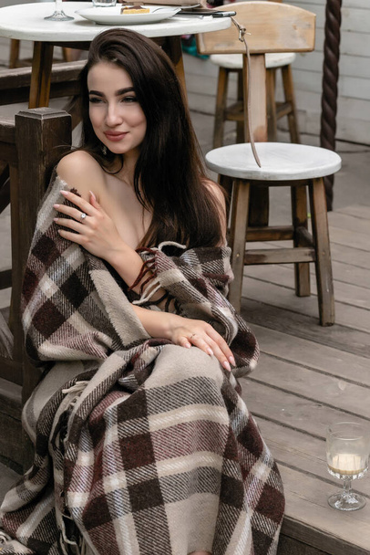 Beautiful girl resting sitting in cafe on the terrace, wrapped in a woolen plaid blanket. Autumn morning. Beautiful young woman with cup of coffee outdoors on autumn day - Valokuva, kuva
