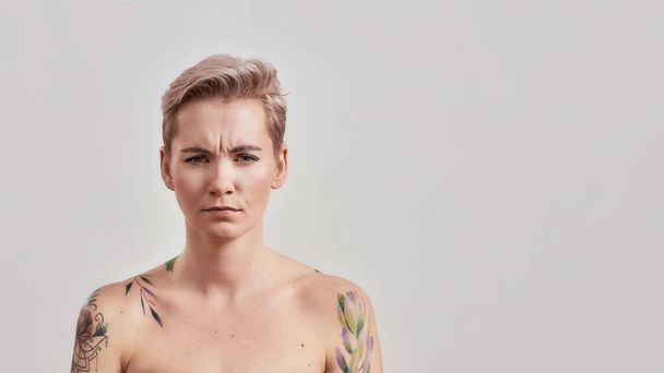 Still kind of miffed. Portrait of dissatisfied half naked tattooed woman with pierced nose and short hair frowning face at camera isolated over light background - Foto, Bild