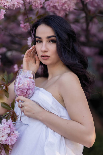 people, rest, comfort and leisure concept - Young woman through the evening streets wrapped in a blanket. Beautiful young girl in the park and a flowering tree sakura holding a glass with petals - Foto, imagen