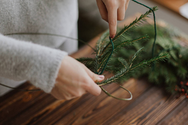 Yeşil köknar dallarını tutan kadınların ellerini kapat ve ahşap masanın arka planında rustik Noel çelengi yap. El yapımı şenlik dekorasyonu, tatil gelişimi. Mevsimlik kış atölyesi - Fotoğraf, Görsel