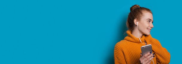 Femme caucasienne avec des taches de rousseur et les cheveux rouges sourit tout en regardant quelque part et tenant son mobile avec des écouteurs sur un mur bleu avec de l'espace libre - Photo, image