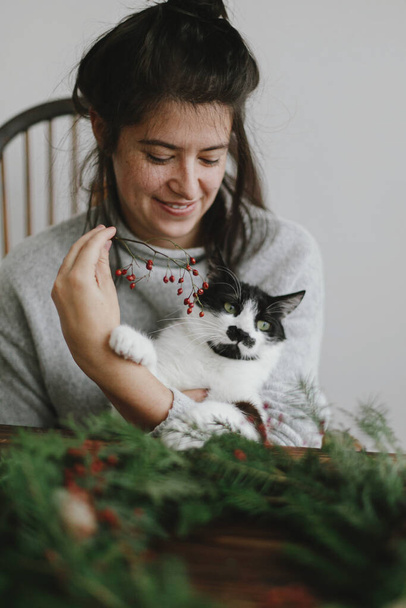 自宅でかわいい猫とクリスマスリースを作る、休日の到来。赤い果実と緑の枝を保持し、愛らしいフェリーネヘルパーとクリスマスリースを作る幸せな女性。スタイリッシュな本物のイメージ - 写真・画像