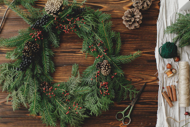 Rustykalny świąteczny wieniec na drewniany stół leżę płaski. Nowoczesny świąteczny wieniec z czerwonymi jagodami, zielonymi gałęziami, szyszkami, nożyczkami, sznurkiem na rustykalnym tle. Warsztaty wakacyjne - Zdjęcie, obraz